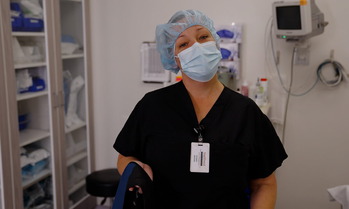 Healthcare worker in black scrubs, surgical cap, and face mask standing in a medical supply room